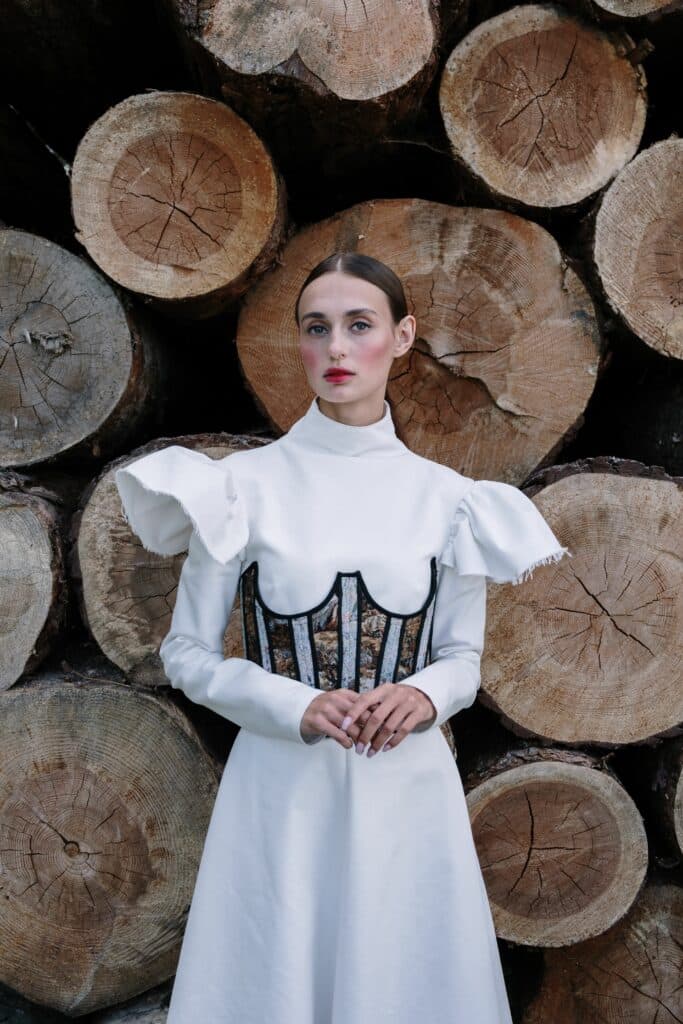 Femme en robe blanche à manches volantées et corset brodé devant un mur de troncs d'arbres empilés