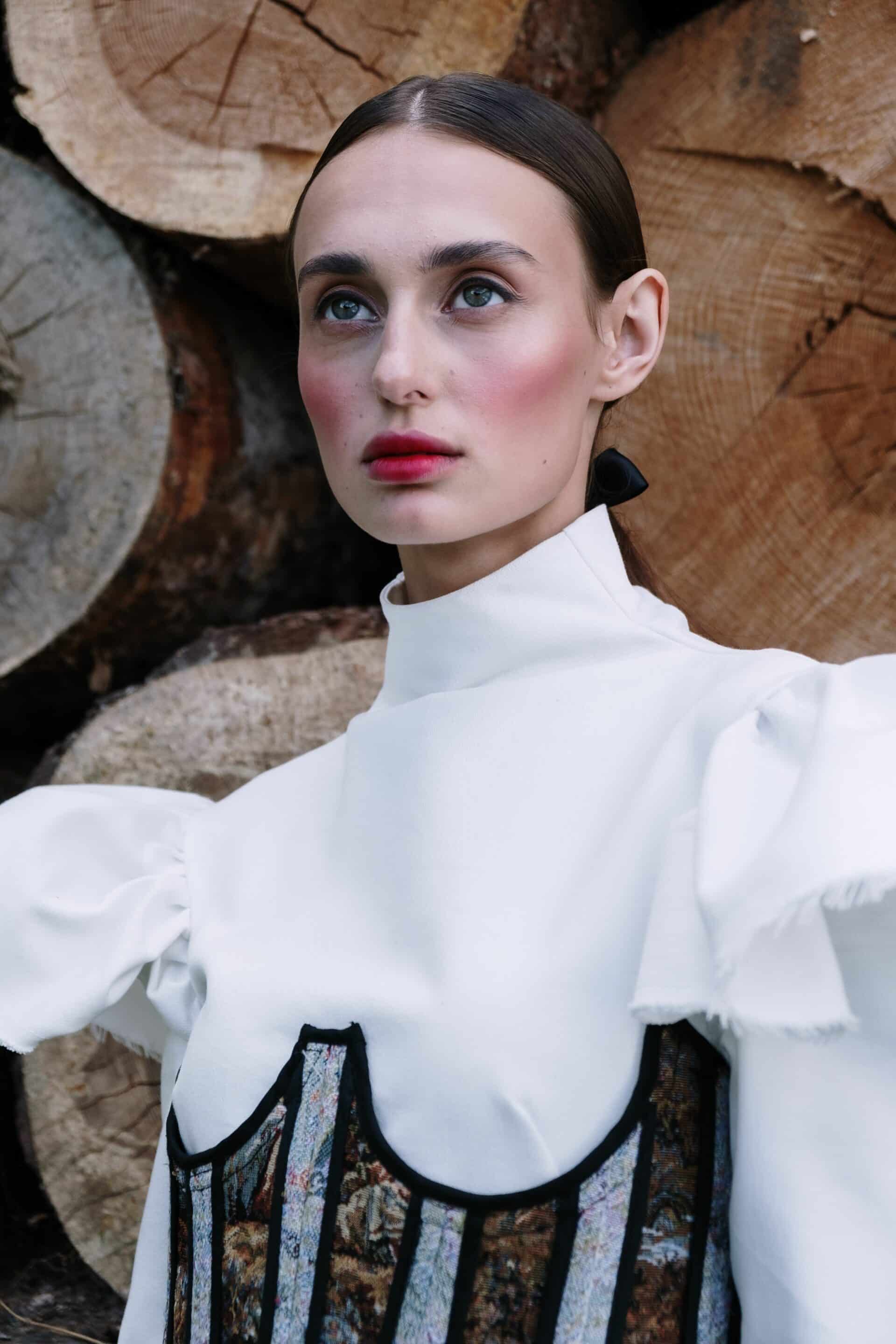 Portrait rapproché d'une femme en haut blanc à col montant et corset brodé, devant des troncs de bois