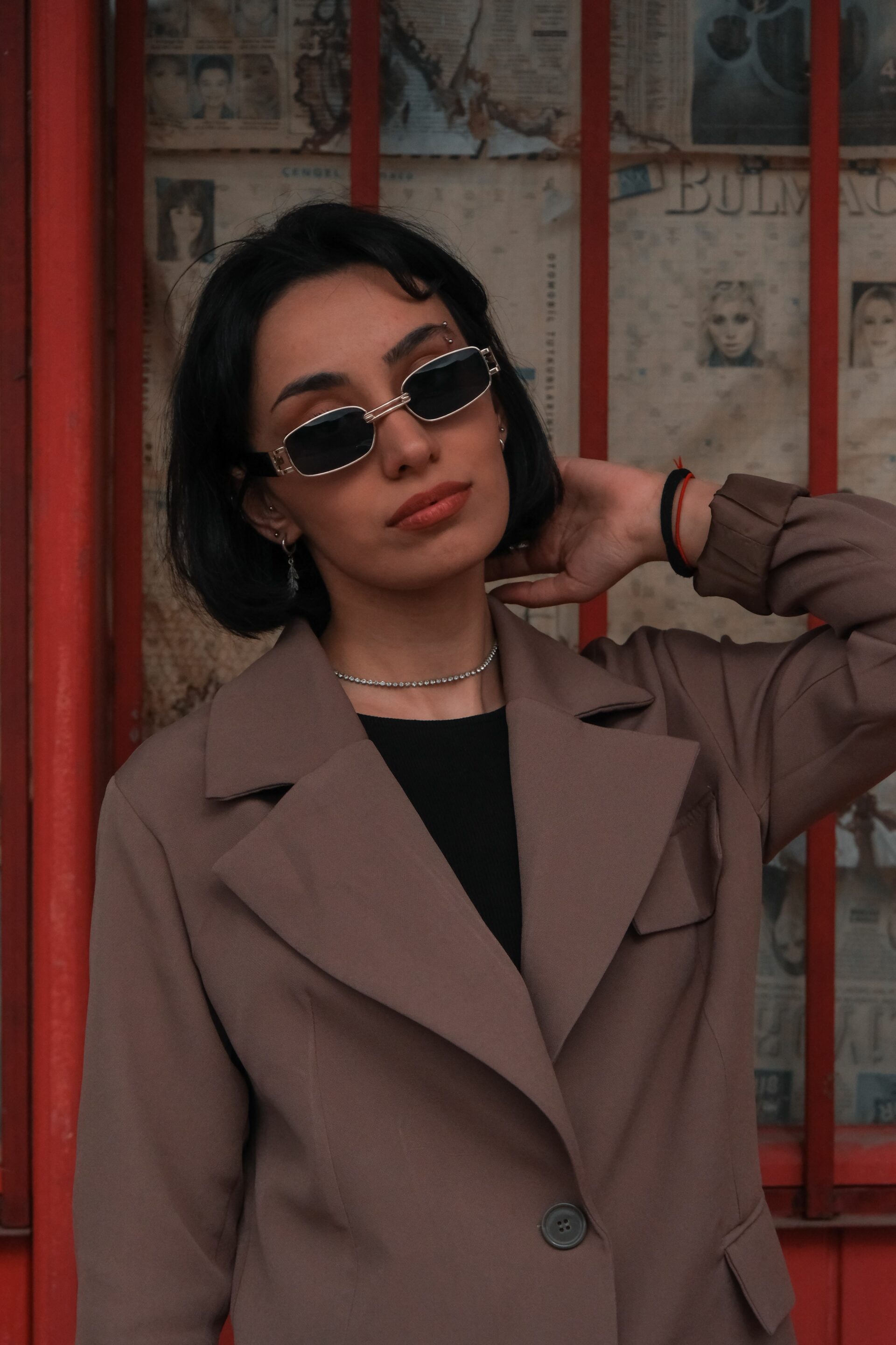 Femme en blazer taupe et lunettes de soleil rectangulaires, la main derrière la nuque, devant une porte rouge