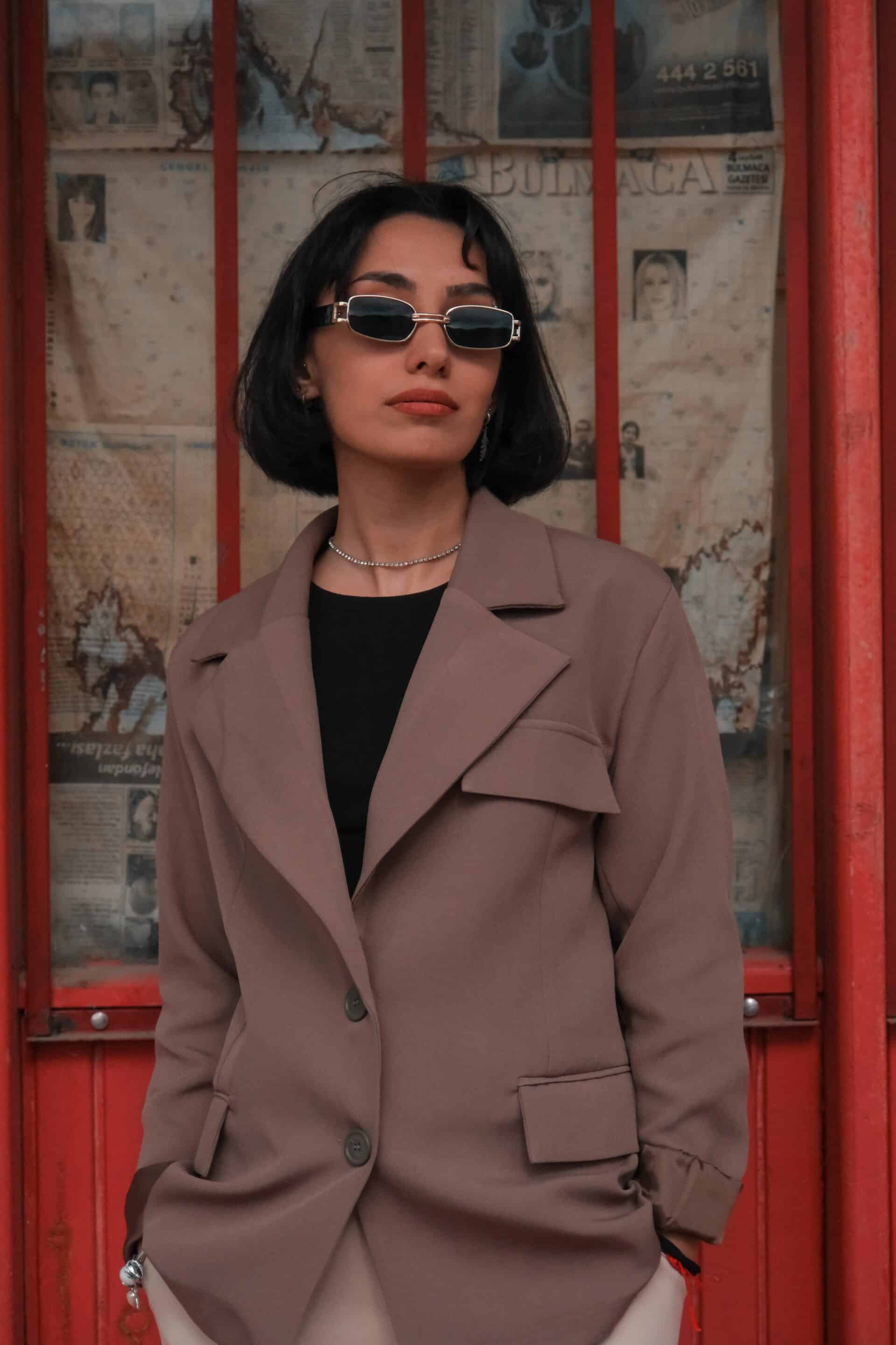 Femme en blazer taupe et lunettes de soleil, mains dans les poches, devant une vitrine rouge tapissée de journaux