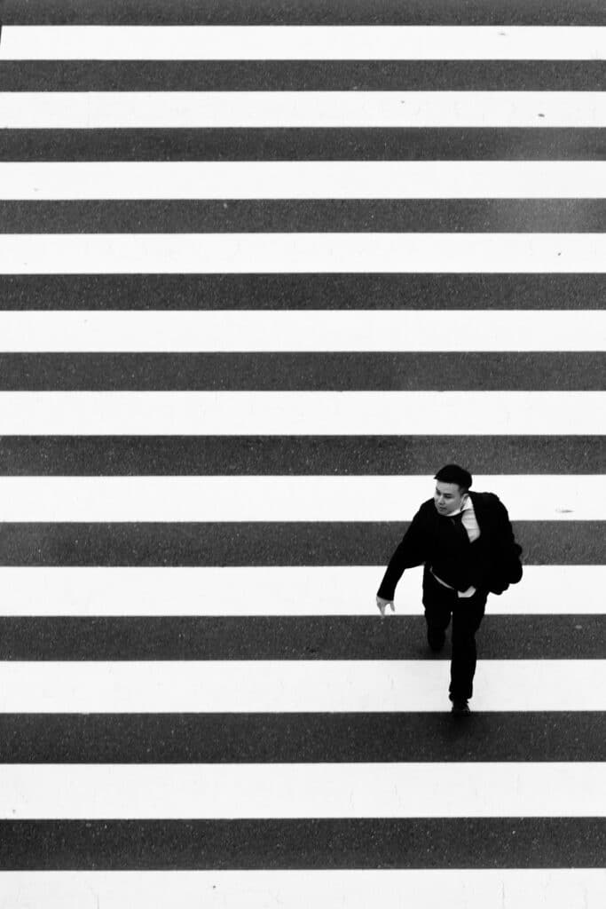 Homme en costume noir traversant un passage piéton rayé, vue du dessus en noir et blanc